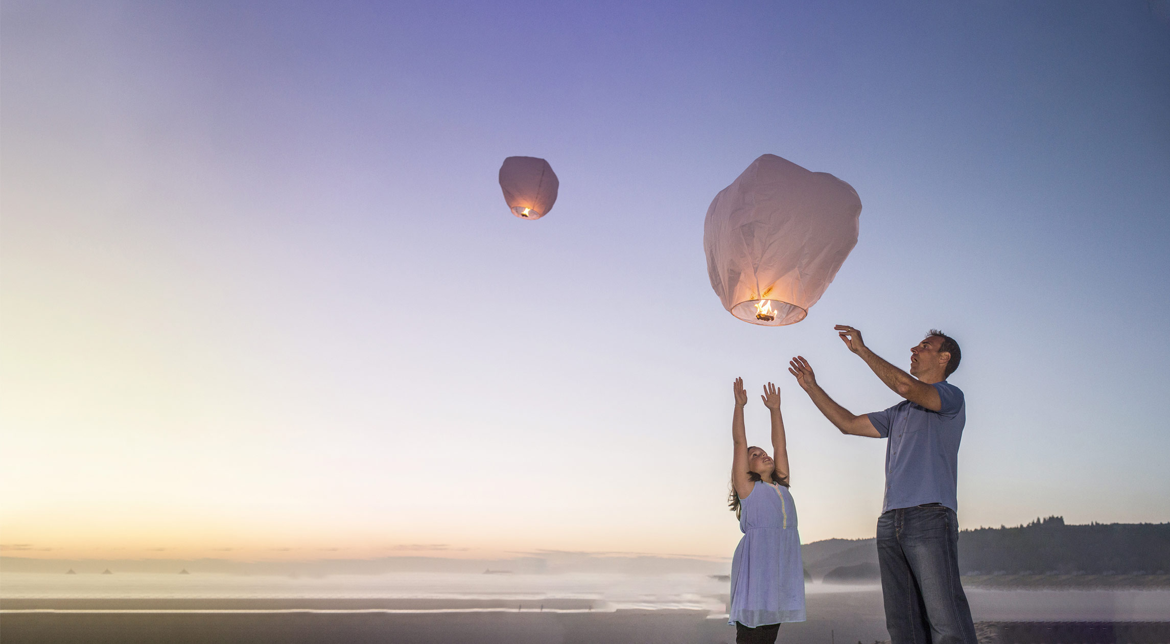 Father and a daughter releasing sky lanterns on a beach at sunset, symbolising hope, dreams, and new beginnings.