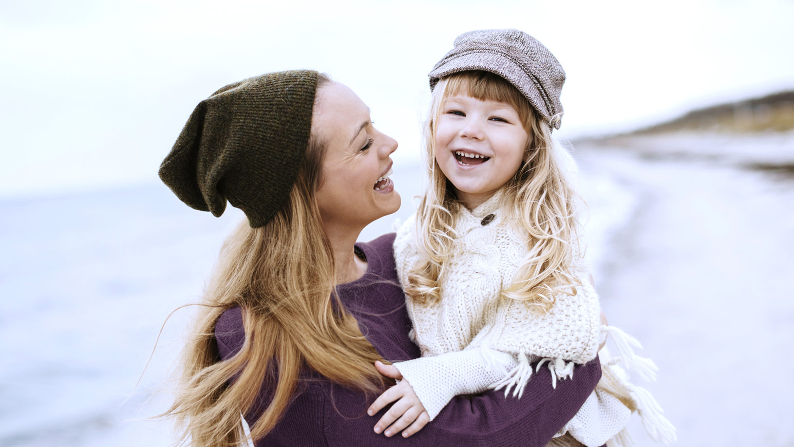Mother and young daughter smiling and embracing outdoors in winter, both wearing warm hats